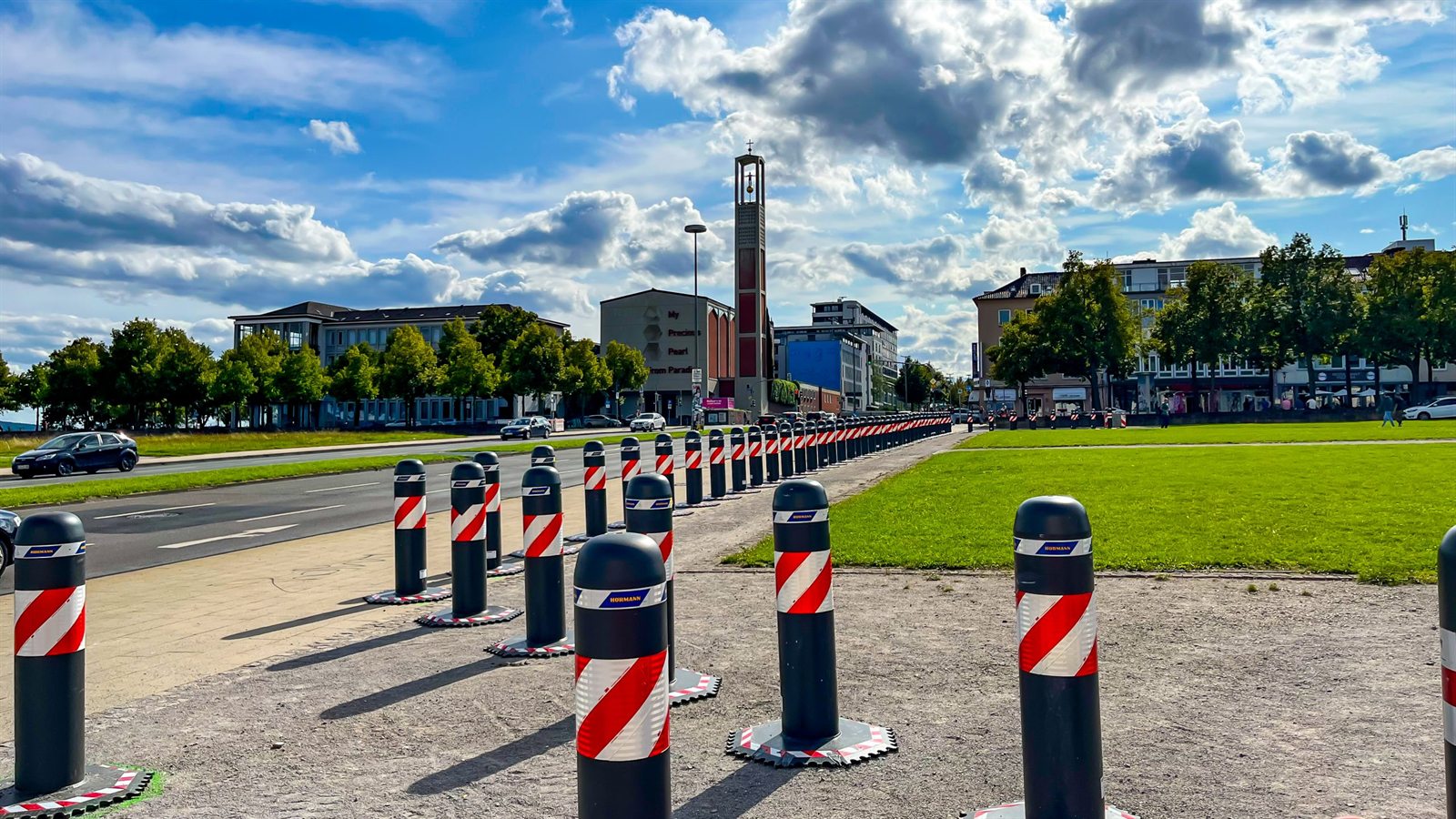 Reihe mobiler Poller mit rot-weißer Kennzeichnung entlang einer Straße, dahinter Kirche und Stadt